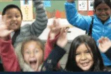 Four children are enthusiastically raising their hands and smiling. They are standing close together, appearing excited and cheerful, with a colorful background behind them.