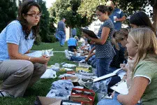 A woman crouches on grass, engaged in discussion with a group of people sitting in a line. They appear to be participating in an outdoor educational activity, using clipboards and various materials spread around them. Trees and a building are in the background.