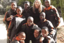 A group of smiling people, including eight children wearing matching black and orange jackets, are gathered around an adult with long hair. They are outdoors, with trees visible in the background.