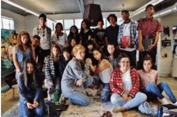 A group of diverse young people sitting and standing indoors, smiling at the camera. They are casually dressed, and the setting appears to be a bright room with art materials in the background.