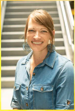 A woman with long brown hair smiles at the camera, standing in front of a staircase. She is wearing a blue button-up shirt and large round earrings.