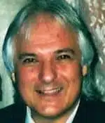 A smiling man with medium-length gray hair, wearing a dark suit, photographed indoors against a patterned background.