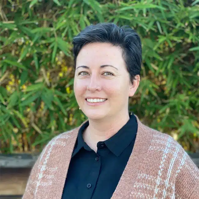 A person with short dark hair smiles while standing in front of green foliage. They are wearing a black shirt and a brown patterned cardigan.