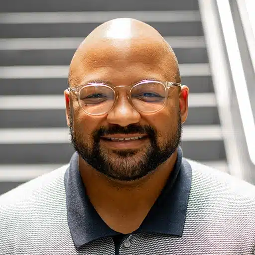 Curtis Taylor, a man with glasses, a short beard, and a shaved head, smiles at the camera in a collared shirt. He stands in front of a staircase with blurred steps in the background.