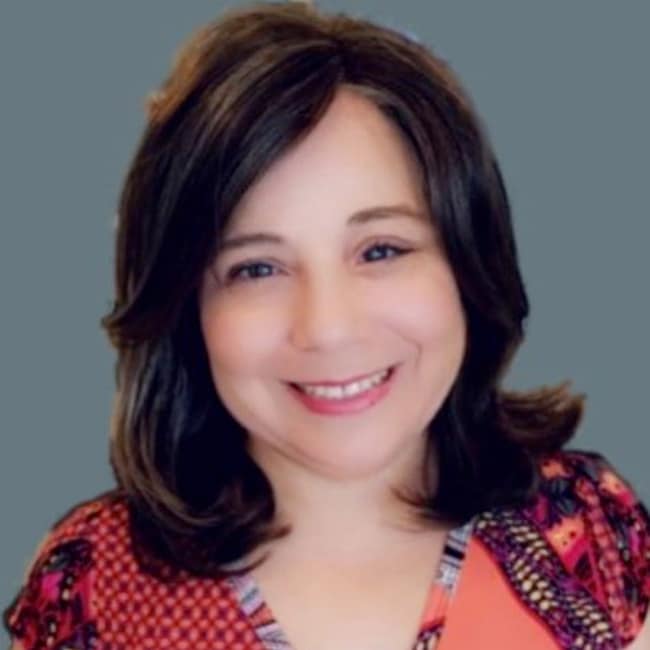A person with shoulder-length brown hair, smiling, exudes confidence in a vibrant red and purple patterned top. Set against a plain gray background, this portrait captures the essence of Zoltan Sardas distinctive style.