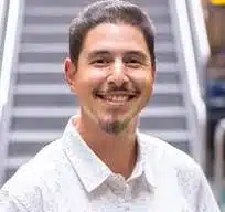 David Garcia, a man with dark hair and a goatee, wearing a white patterned shirt, smiles in front of a blurred staircase background.