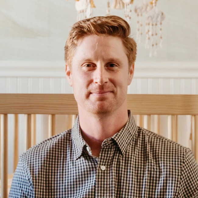 A man with short, light brown hair and a checked shirt is smiling gently while sitting in front of a wooden crib and a light-colored wall with hanging decorations.