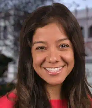 Smiling woman with long brown hair wearing a red top, standing outdoors with a blurred background.