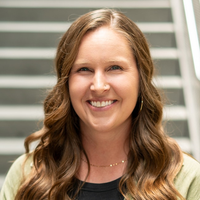 Smiling woman with long, wavy brown hair stands in front of gray stairs. She is wearing a green cardigan over a black top and small hoop earrings.