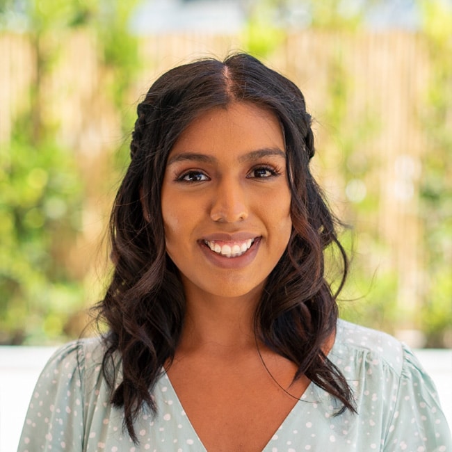 Esmeralda Garcia, with long, wavy dark hair and a light green polka dot top, smiles at the camera outdoors, framed by a blurred background of greenery.