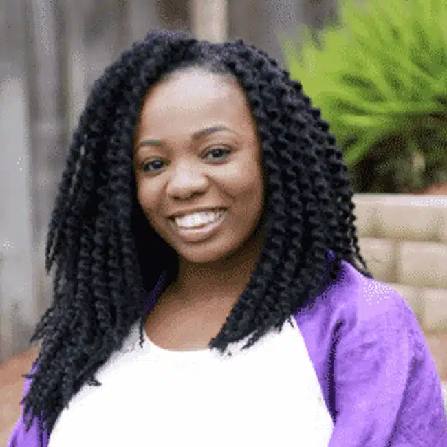 A person with long, curly hair smiles at the camera. They wear a white top and a purple jacket. The background includes green plants and a wall.