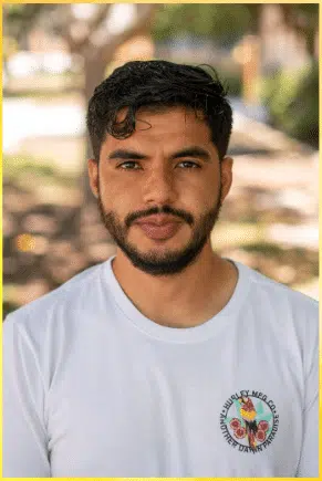 A person with short black hair and a beard stands outdoors, wearing a white t-shirt with a colorful logo on the left chest. The background is blurred, showing trees and sunlight.