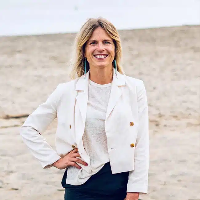 A woman stands on a beach, smiling. She is wearing a white blazer over a light top and has long earrings. The background shows sandy terrain under a cloudy sky.