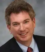 A man with curly brown hair is smiling at the camera. He is wearing a dark suit, white shirt, and red patterned tie. The background is a soft, out-of-focus gray.