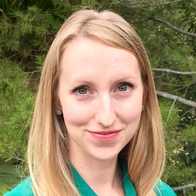 A woman with long blonde hair and a green top smiles in an outdoor setting with greenery in the background.