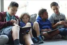 Four children sit closely together near a window, reading books and interacting with each other. They appear engaged and relaxed, with natural light streaming in beside them. They are casually dressed.