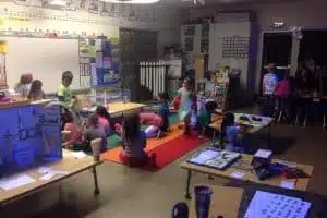 A classroom with young children engaged in various activities. Some sit on colorful mats, while others work at tables filled with books and educational materials. Walls are decorated with charts and artwork. Lighting is dim.
