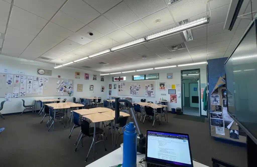 A classroom with several hexagonal tables and chairs, a wall decorated with posters and photos, and a laptop and water bottle on a desk in the foreground. Fluorescent lights illuminate the room, and theres a clock on the wall.