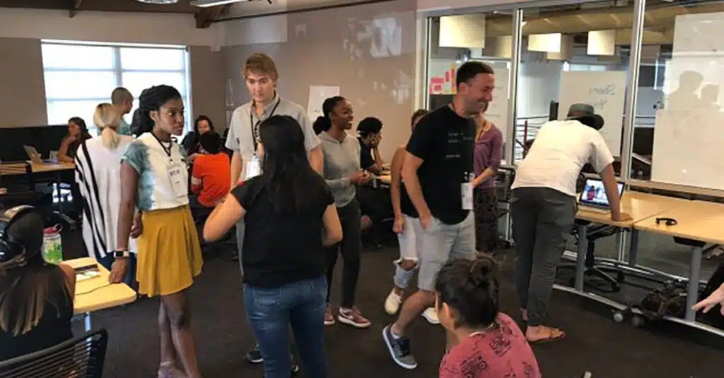A group of people standing and sitting in a modern office space, conversing and working at desks. Large windows and glass partition walls create an open and collaborative atmosphere.
