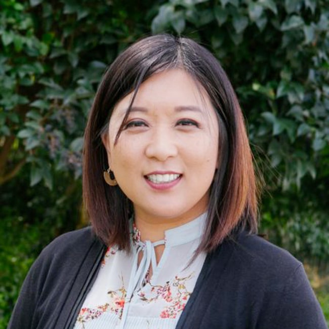 A person with shoulder-length hair smiles warmly. They are wearing a floral blouse and a black cardigan. The background is filled with lush green leaves.