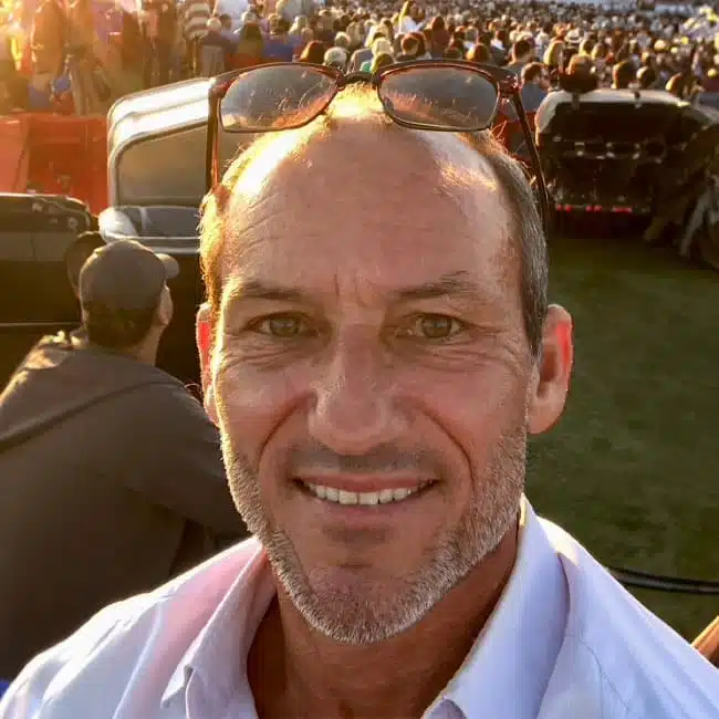 A man with short hair and stubble, wearing glasses on his head and a white shirt, smiles at the camera. He is outdoors at an event with a large crowd behind him, captured in the warm glow of sunset.