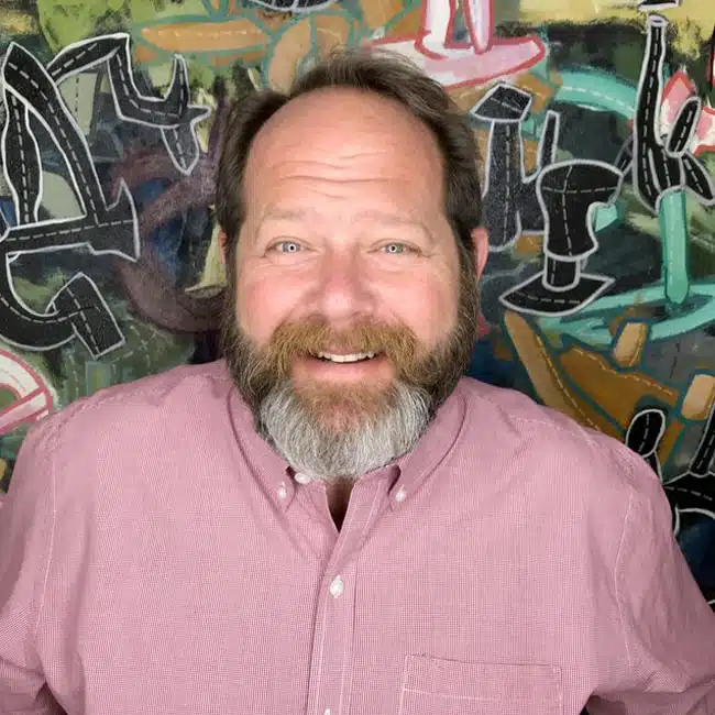 A bearded man wearing a pink shirt smiles warmly in front of a colorful, abstract mural, embodying the vibrant spirit nurtured at the High Tech High Graduate School of Education.