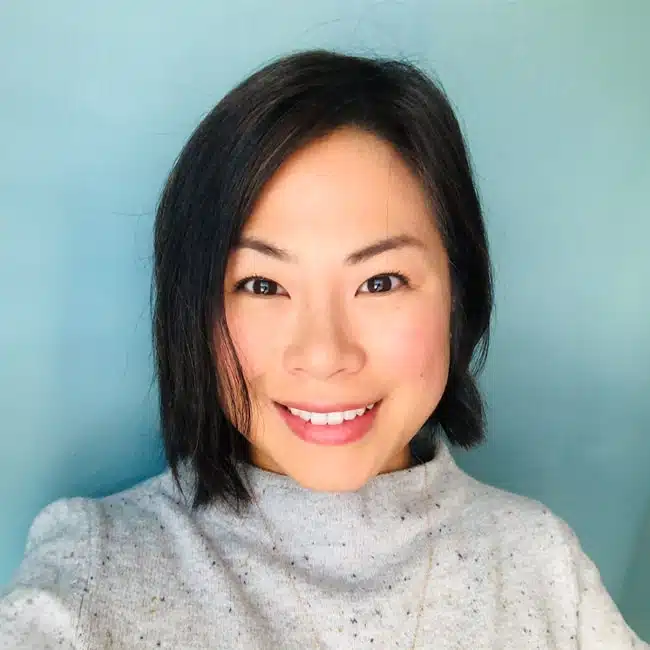 A person with shoulder-length black hair smiles at the camera. They are wearing a light gray sweater against a light blue background.