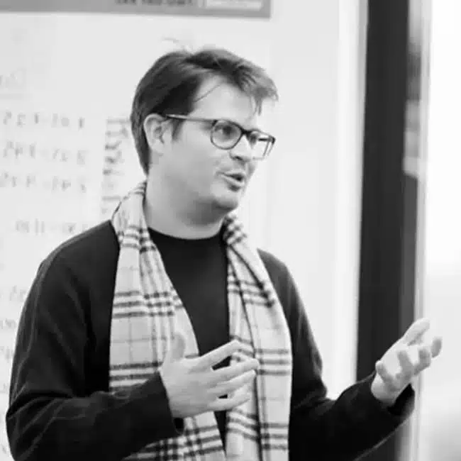 Man with glasses and a plaid scarf gestures while speaking in a classroom. A board with notes is visible in the background. Black and white image.