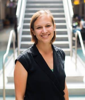 A person with short hair smiles while standing in front of a staircase. They are wearing a black shirt in a brightly lit indoor setting.
