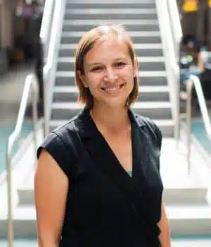 A person with short hair smiles while standing in front of a staircase. They are wearing a black shirt in a brightly lit indoor setting.