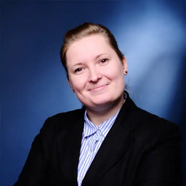 Juliane Fricke, with light brown hair, is smiling at the camera. They are wearing a black blazer over a blue and white striped shirt. The background is a gradient of dark to light blue.