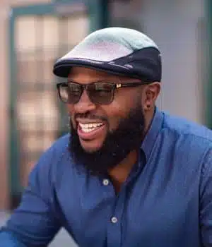 A smiling man with a beard, wearing glasses, a flat cap, and a blue button-up shirt. He is outdoors, with blurred background of windows and walls.
