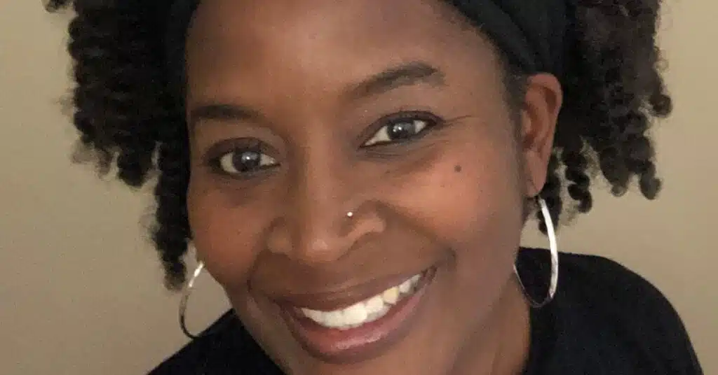 A person with curly hair and hoop earrings smiles at the camera. They are wearing a black top and a headband.