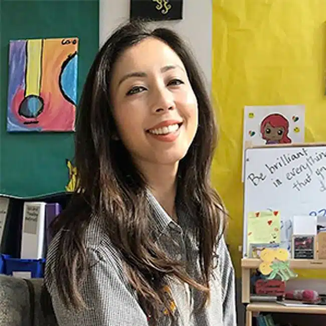 A woman with long hair smiles while sitting in a room filled with colorful artwork and positive messages. Behind her is a painting of a guitar and a poster that reads, Be brilliant in everything that you do.