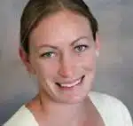 A person with light-colored hair smiling at the camera against a neutral background. They are wearing a light-colored top.