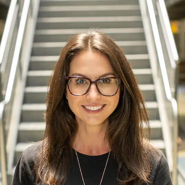 A person with long brown hair and glasses smiles in front of a blurred staircase background, wearing a black top.