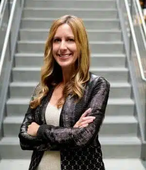 A person with long, light brown hair stands confidently with arms crossed, wearing a black leather jacket over a white top. They are standing in front of a staircase, smiling at the camera.