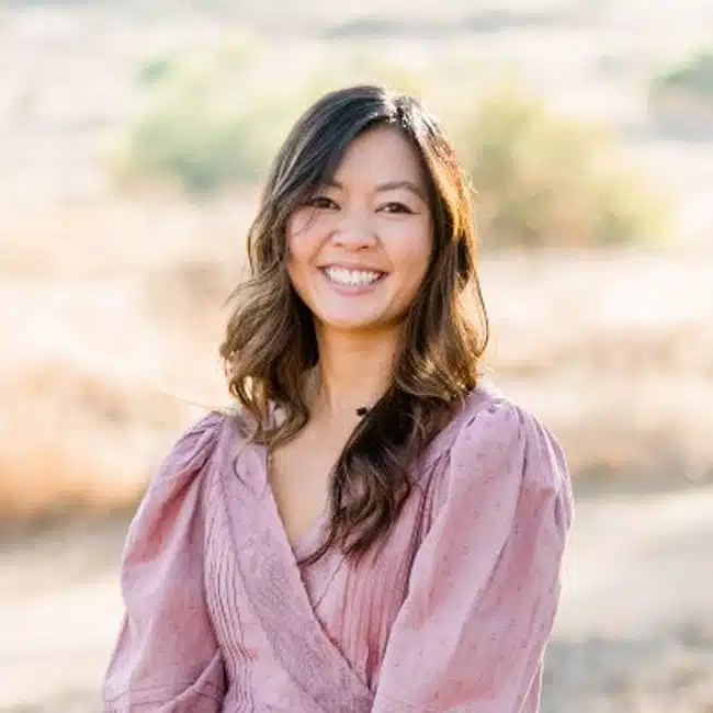 A woman with long wavy hair smiles warmly. She is wearing a pink blouse with puffed sleeves. The background shows an outdoor landscape with soft, blurred greenery and earth tones.