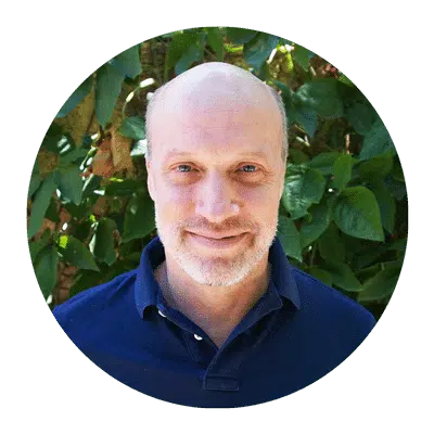 A person with a bald head and beard is smiling, wearing a navy blue shirt. Lush green leaves form the background.