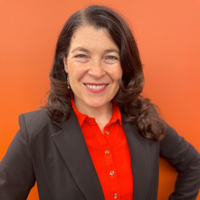 A woman with long dark hair smiles at the camera. She is wearing a black blazer over a bright orange shirt, standing in front of an orange background.