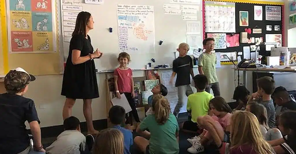 A teacher stands at the front of a classroom, engaging a group of young students. Some students are seated on the floor, while a few stand near the whiteboard. The room is colorful with various educational posters on the walls.