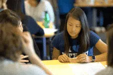 A young woman in a blue shirt is focused on paperwork at a table with others. The setting appears to be a classroom or workshop, with participants engaging in discussions and activities.