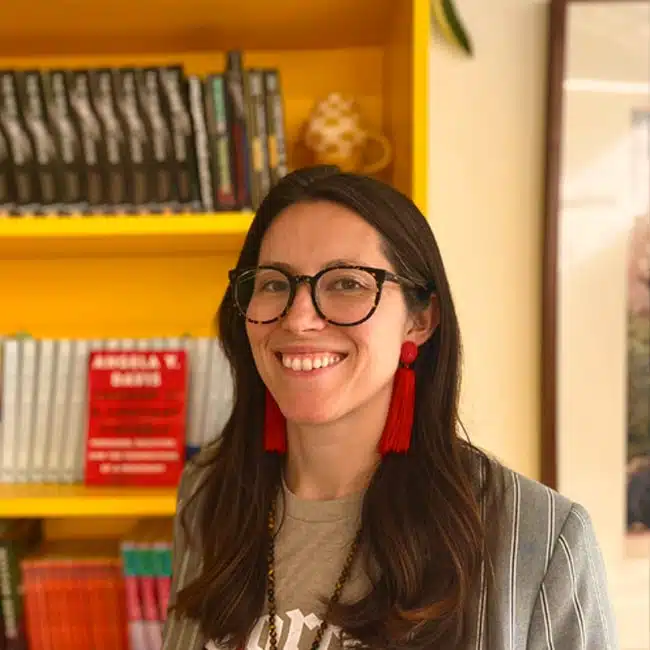 A person with long dark hair, wearing glasses and red earrings, smiles in front of a yellow bookshelf filled with books. They are dressed in a striped jacket and a t-shirt.