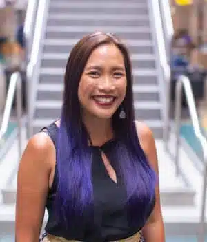 A smiling woman with long, straight hair partially colored purple stands in front of a staircase, wearing a black sleeveless top and earrings. The background is blurred, highlighting her cheerful expression.