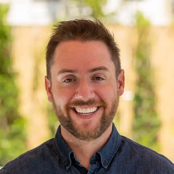 A smiling man with short hair and a beard is wearing a dark button-up shirt, perfect for his Instagram profile. The background is blurred, showing greenery and a light-colored surface, creating an ideal alt text description for accessibility.