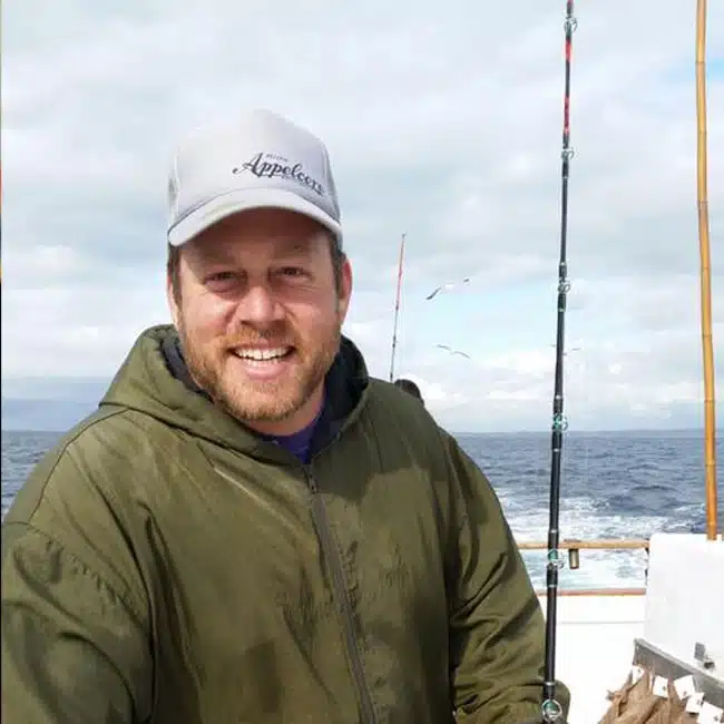 A smiling person in a green jacket and gray cap is on a boat, holding a fishing rod. This Instagram-worthy image captures the ocean and a few birds visible in the background under a cloudy sky.