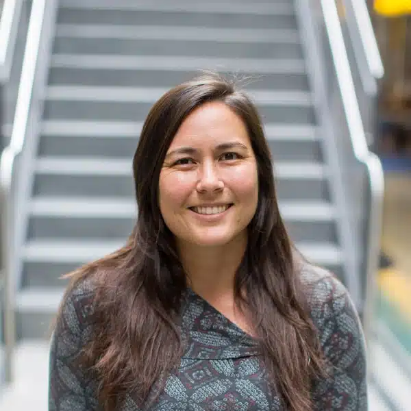 A person with long brown hair and a patterned top smiles while standing in front of a staircase, capturing an image perfect for sharing on Facebook.