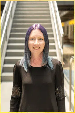 A person with purple and blue hair, wearing a black long-sleeve top, stands in front of a staircase. They are smiling and the background is softly lit.