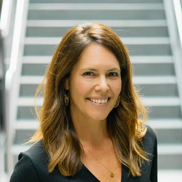 A woman with long brown hair is smiling at the camera. She is wearing a black top and gold earrings, with a pendant necklace. Behind her is a blurred staircase.