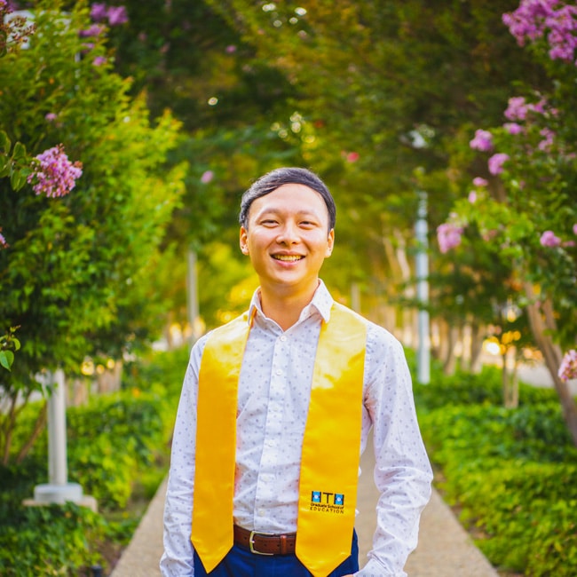 Michael Chin, a young person in a white shirt and yellow graduation stole, stands smiling on a garden path lined with green trees and pink flowers, sunlight streaming through the leaves.
